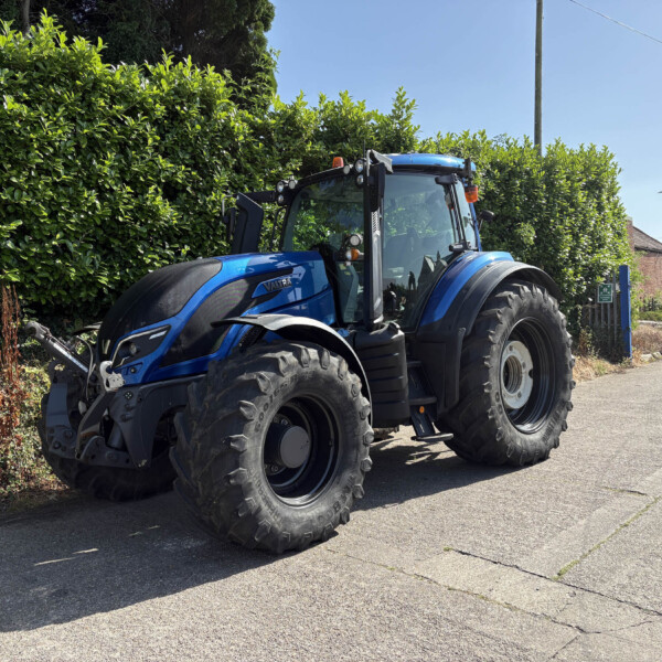 Blue tractor parked along a street beside a tall green hedge on a sunny day under a clear blue sky.