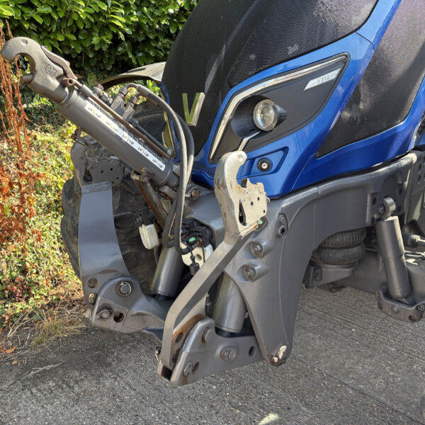 Close-up of a blue tractor’s front end showing the hydraulic lift arms and exposed metal components, with greenery in the background.