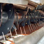 Close-up of a rusted, curved metal blade assembly inside a dark metal housing, with wet surfaces and several thin rods crossing the scene.