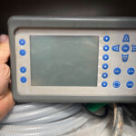Hand holding a grey handheld diagnostic/controller device with a screen and blue circular and triangular buttons on the right side.