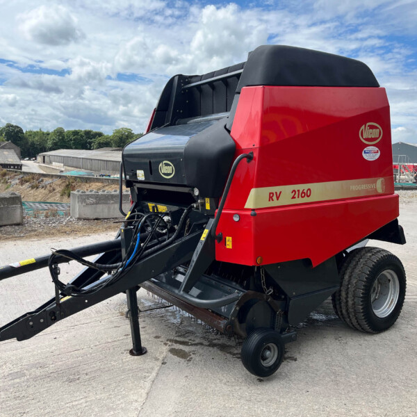 Red Vicom RV 2160 agricultural header parked in an outdoor yard with warehouses in the background, showing machinery and hoses nearby.