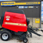 Red Vicon RV 2160 baler in the foreground at a New Holland Agriculture dealership, with a dark gray building in the background and blue sky above.