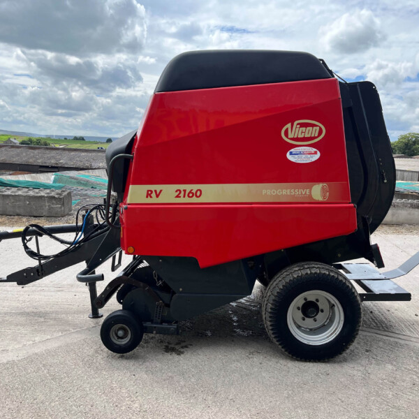 Red Vicon RV 2160 agricultural baler on a concrete surface with a cloudy sky and rural buildings in the background