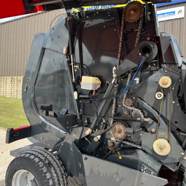 Close-up of exposed drive chain, gears, and wiring inside a gray agricultural harvester, outdoors near a building with signs visible in the background.