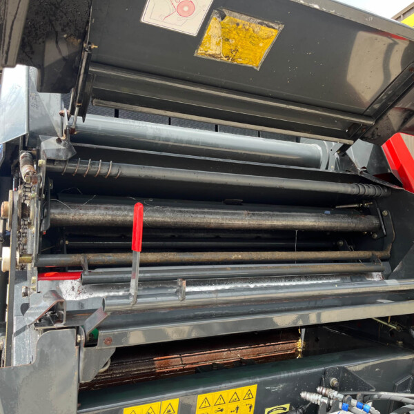 Close-up of a large industrial machine's metal framework with rollers, guards, and red handles amid dark gray panels and yellow warning decals at a worksite.