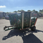 Green agricultural machinery on a trailer in an outdoor yard with a metal fence, parked cars, and a red waste bin nearby.