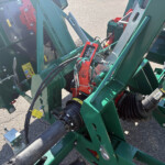 Close-up of a green agricultural machine frame with hoses, bolts, and warning labels nearby.