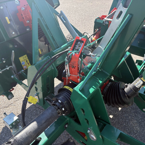 Close-up of a green agricultural machine frame with hoses, bolts, and warning labels nearby.
