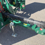 Close-up of a green agricultural machine showing hydraulic hoses, a black protective shaft, and metal brackets on an asphalt surface; yellow safety decals are visible.