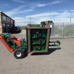 Green and red agricultural equipment in a parking area outside a brick and metal industrial building, with a tall corrugated fence and blue sky above.