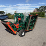 Green and red agricultural implement with wheels parked in a lot, background shows a metal fence and houses under a blue sky.
