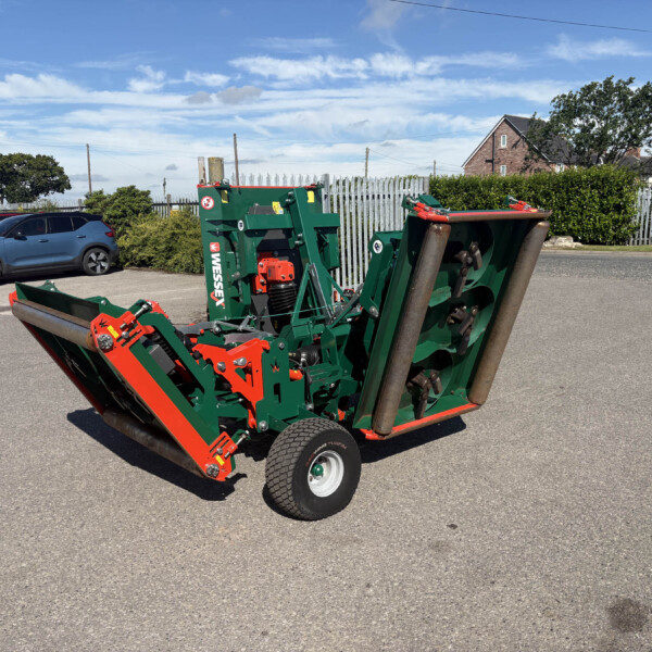 Green and red agricultural implement with wheels parked in a lot, background shows a metal fence and houses under a blue sky.