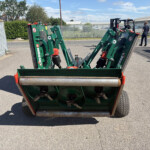 Rear view of a green farming implement with rollers on an asphalt lot; two workers near a metal fence in the background