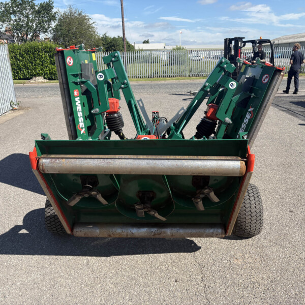 Rear view of a green farming implement with rollers on an asphalt lot; two workers near a metal fence in the background