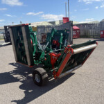 Green agricultural implement attached to a trailer leans on its side in an outdoor yard, with a tall metal fence, red waste bins, and blue sky overhead.