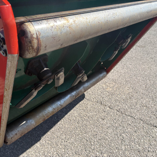 Close-up of a rusted metal roller on a green agricultural machine with a red support frame and bolts visible on a paved surface.