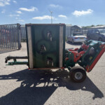 Overturned green agricultural equipment in a parking lot beside a tall metal fence, with cars in the background and a blue sky above.