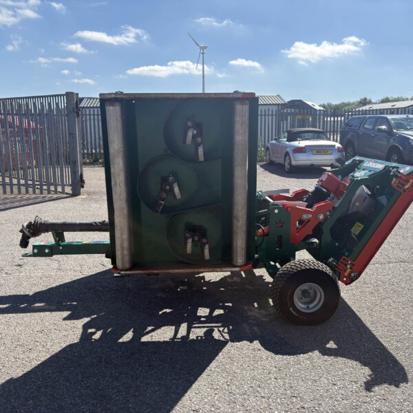 Overturned green agricultural equipment in a parking lot beside a tall metal fence, with cars in the background and a blue sky above.