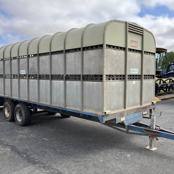 Large metal livestock trailer on a wheeled trailer, with a blue tractor visible in the background and a cloudy sky above.