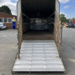 Open flatbed trailer with metal diamond-plate ramp at an industrial yard, metal sides, and blue sky above.