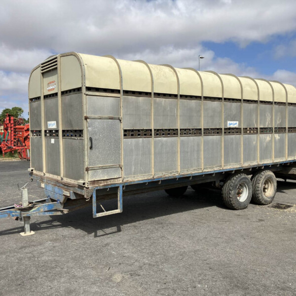 Large metal farm trailer on a wheeled trailer in a yard with red machinery in the background and a cloudy sky above.