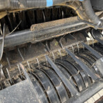 Close-up of dusty metal blades and tines inside an agricultural harvester header with straw debris and wiring nearby.