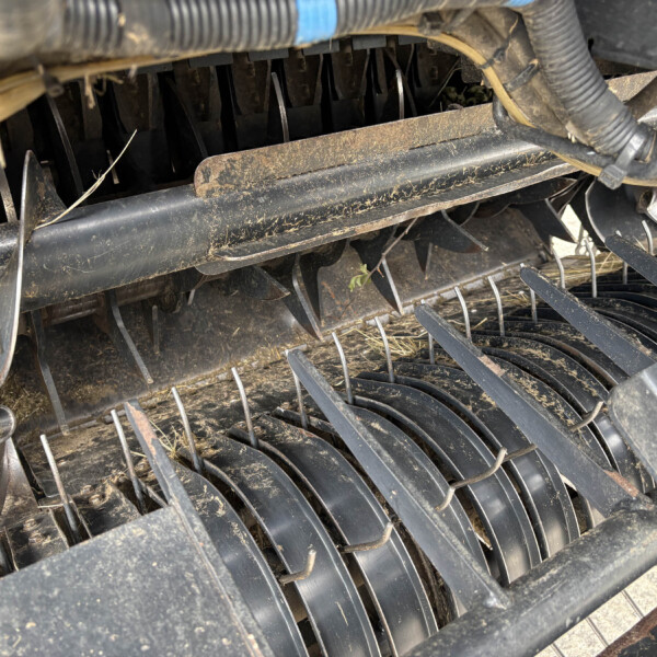 Close-up of dusty metal blades and tines inside an agricultural harvester header with straw debris and wiring nearby.