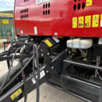 Red agricultural machine with numerous yellow warning stickers and tangled hydraulic hoses at a farm yard.