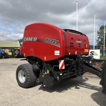 Red Case IH RB455 round baler on a dealership lot, with blue tractors and a cloudy sky in the background.