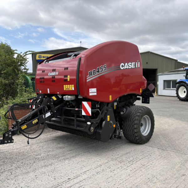 Red CASE IH RB455 baler in a farm yard with a blue tractor nearby and a gray industrial building in the background.