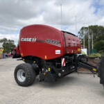 Red CASE IH RB455 baler parked in an outdoor lot with other heavy machinery and a cloudy sky behind it, showing its rounded body and large tires.