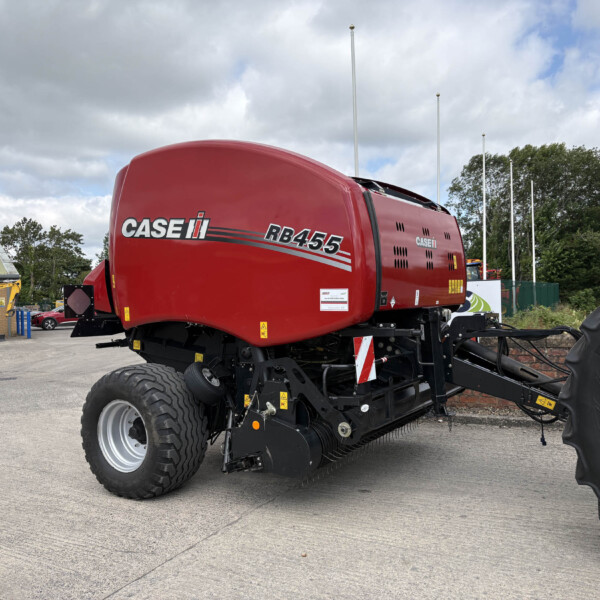 Red CASE IH RB455 baler parked in an outdoor lot with other heavy machinery and a cloudy sky behind it, showing its rounded body and large tires.