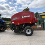 Red Case IH RB455 agricultural header on a tractor at a dealership, with a blue tractor nearby and a New Holland sign in the background, outdoor lot scene.