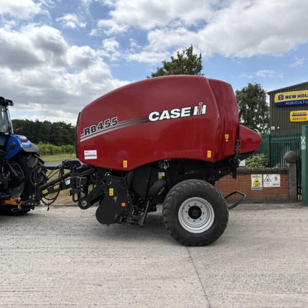 Red Case IH RB455 agricultural header on a tractor at a dealership, with a blue tractor nearby and a New Holland sign in the background, outdoor lot scene.