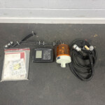 Automotive control panel with display, orange beacon, mounting hardware, and tangled cables on a concrete floor surface.