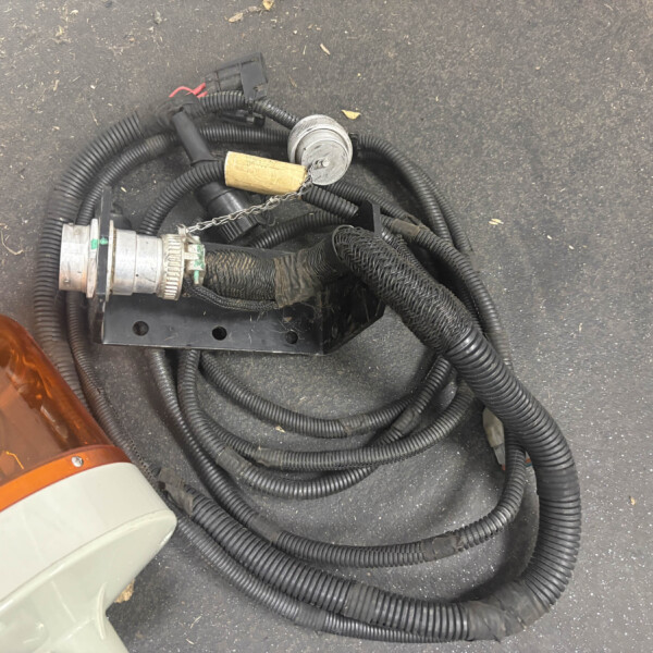 Wiring harness with black corrugated sleeves and a round metal connector, on asphalt beside an orange vehicle light.