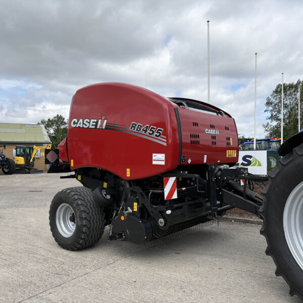 Red CASE IH RB455 baler in a farm equipment yard, with tractors and a brick building under a cloudy sky.