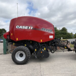 Close-up of a red Case IH RB455 baler attached to a blue tractor, with other tractors visible in the background.