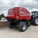 Red CASE IH RB455 baler hitched to a blue tractor, parked by a brick wall under a cloudy sky with power lines in the background.
