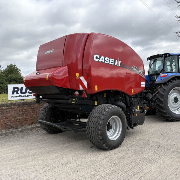 Red CASE IH RB455 baler hitched to a blue tractor, parked by a brick wall under a cloudy sky with power lines in the background.