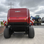 Rear view of a red CASE IH agricultural machine on a flatbed trailer at a transport yard with other equipment nearby under a cloudy sky.