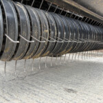 Close-up of a dusty cylindrical metal rake/spine with curved rings and evenly spaced tines on an agricultural machine.