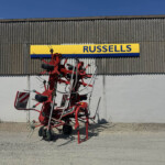 Red agricultural machinery stacked outside against a ribbed metal wall beneath a yellow Russell sign against a blue sky.