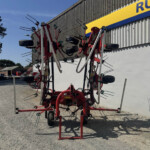 Large red hay rake with many metal tines on a gravel lot beside a corrugated wall building, outdoor farm equipment.