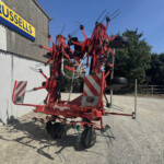 Red agricultural cultivator with multiple tines and wheels parked outside a building on a gravel lot.