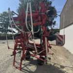 Red multi-row agricultural cultivator with tines and wheels, parked on gravel beside a white-wall building and trees in the background.