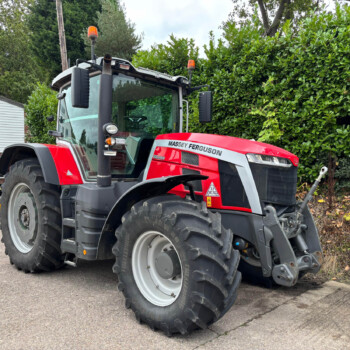 Red Massey Ferguson tractor with large front tires parked in a lot, hedge and sign in the background.