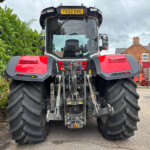 Front view of a large red tractor with oversized tires, outdoors beside a hedge and brick houses.