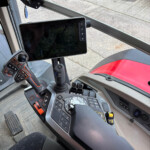 Interior view of a heavy machinery cab with a large touchscreen monitor mounted to a bar, a control joystick, and a complex keypad at the console, outside showing orange attachment equipment.