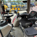Inside a tractor cab: steering wheel and control panel with screens and levers, facing a yard filled with blue and yellow tractors parked outside the front windshield.